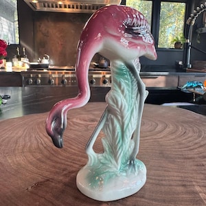 May include: A ceramic flamingo figurine featuring a glossy finish with a deep pink head and neck, a white body, and light green foliage detailing at the base. The bird is depicted in a downward-facing posture with its head curved toward the ground.