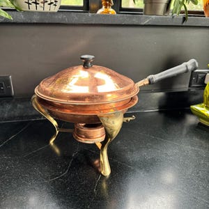Peut inclure: Un plat à réchaud en cuivre avec un couvercle assorti, doté d'une poignée noire et d'un bouton noir sur le couvercle. Le plat est soutenu par trois pieds en laiton incurvés et comprend un brûleur central sous la cuve pour le chauffage.