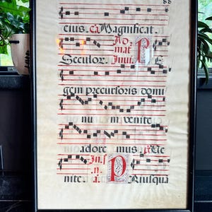 May include: A framed manuscript page featuring black square musical notation on red staff lines, with black gothic calligraphy text and ornate red and white illuminated capital letters. The page is numbered 88 in the top right corner.