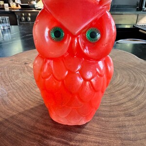 May include: A bright red owl-shaped candle featuring a molded feather texture across its body and two large, round, vibrant green eyes. The wax figure has pointed ear tufts at the top of its head and a smooth, glossy finish throughout its surface.
