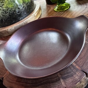 May include: A dark brown, circular ceramic dish with a smooth, matte finish and two integrated side handles. The dish features a shallow, wide basin and a slightly raised rim, presenting a uniform, deep earth-toned color throughout its surface.