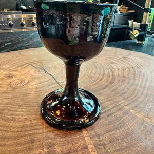 May include: A dark brown ceramic goblet with a glossy finish and a wide, rounded bowl. The chalice features a thick, sturdy stem and a circular base, with subtle hints of green glaze visible along the upper rim of the vessel.
