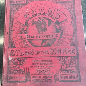 Op de afbeelding: Een rood gebonden boek getiteld Cram's Ideal Reference Atlas of the World, gedateerd 1902. De kaft bevat een illustratie van een wereldbol, de tekst With the Complete 1900 Census by Counties and Towns, en de uitgever Geo. F. Cram, Chicago & New York.