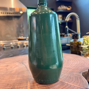 Könnte beinhalten: Eine dunkelgrüne Glasflasche mit einem glatten, zylindrischen Körper und einem schmalen Hals. Am Boden der Flasche befindet sich ein geprägter Schriftzug mit der Aufschrift FONSECA. Die Oberfläche hat ein glänzendes Finish und reflektiert Licht.
