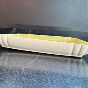 May include: A rectangular ceramic serving dish featuring a smooth cream-colored exterior and a contrasting pale yellow interior. The vessel has a shallow depth with vertical fluted detailing along the sides and rounded corners.
