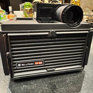 May include: A black Bell & Howell AF 66 slide projector featuring a horizontal slatted front panel, a top-mounted lens with a 100mm f/3.5 specification, and a rectangular brand logo plate on the front with the text Bell & Howell AF 66.