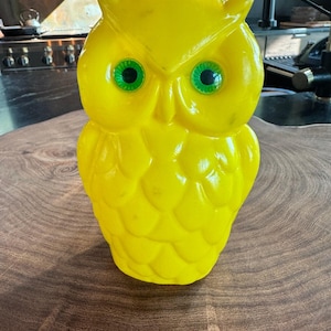 May include: A bright yellow plastic owl figurine featuring prominent, textured feather details across its body and two large, round, vibrant green eyes with black pupils. The owl has pointed ear tufts at the top of its head and a smooth, molded beak.