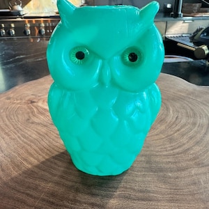May include: A bright turquoise owl-shaped candle featuring a molded feather texture across its body. The owl has two prominent, pointed ear tufts at the top and two small, dark, circular eyes set within the smooth, rounded facial disc area.