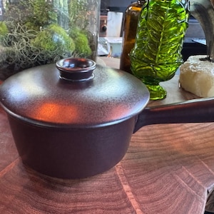 Puede incluir: Un cazo de cerámica de color marrón oscuro con tapa a juego y un mango largo. La pieza presenta un acabado mate liso con una textura ligeramente moteada en toda la superficie. La tapa incluye un pequeño pomo central para facilitar su uso.