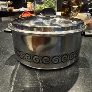 May include: A round, stainless steel cooking pot with a matching lid. The pot features a black, repeating spiral pattern encircling the lower exterior. It has a black handle on the lid and black side handles, presenting a polished, reflective metallic finish.