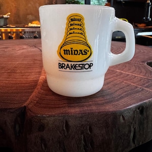 Puede incluir: Una taza de vidrio lechoso blanco con un gráfico amarillo de un diseño de silenciador apilado, con el texto Midas y la palabra BRAKESTOP impresa en negro debajo. La taza tiene un asa en forma de D y un diseño de base escalonada.
