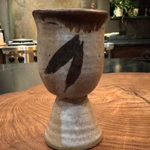 May include: A ceramic goblet with a speckled beige and tan finish, featuring a dark brown rim and a hand-painted dark brown leaf motif on the front. The vessel has a wide, rounded bowl that tapers down to a sturdy, flared pedestal base.
