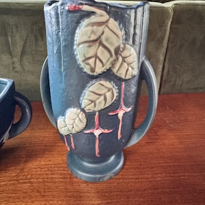 May include: A dark blue ceramic vase with two side handles and a pedestal base. The exterior features raised relief decorations of light green leaves and pink fuchsia flowers. The surface has a textured, matte finish throughout the entire piece.