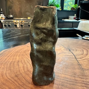 Puede incluir: Un jarrón de cerámica alto y cilíndrico con una textura de superficie irregular moldeada a mano. El jarrón presenta un esmalte de color verde oliva oscuro y moteado con variaciones sutiles de tono y un borde sin esmaltar en la abertura.