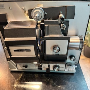 May include: A vintage 8mm Super 8 film projector featuring a metallic silver and black housing. The front panel displays the text 8MM SUPER 8, along with various control knobs, switches for motor and lamp settings, and a speed adjustment dial.