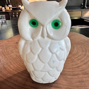 May include: A white owl figurine featuring a molded feather pattern across its body and head. The owl has two prominent, circular, bright green eyes with black pupils, and pointed ear tufts at the top of its head, creating a distinct avian shape.