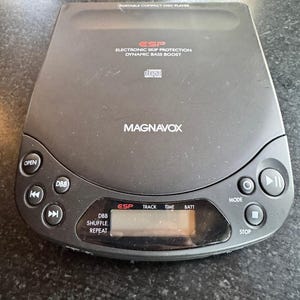 Pode incluir: Um leitor de discos compactos portátil Magnavox preto com proteção eletrónica contra saltos e reforço dinâmico de graves. O dispositivo inclui um ecrã digital e botões de controlo para Open, DBB, navegação, Mode, Play/Pause e Stop.