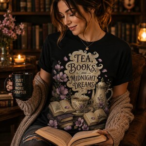 Puede incluir: Una camiseta negra con un estampado gráfico con el texto Tea, Books and Midnight Dreams, rodeado de ilustraciones de una taza de té humeante, una tetera, libros abiertos y flores moradas. Una taza negra con el texto Just One More Chapter.