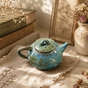 May include: A round ceramic teapot featuring a mottled blue and brown glaze with a glossy finish. The teapot has a curved handle, a short spout, and a matching lid with a small knob handle. The surface displays a textured, speckled appearance.