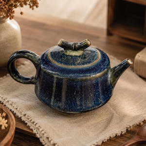 May include: A ceramic teapot with a deep blue, speckled glaze and a glossy finish. The teapot features a rounded body, a curved handle, a short spout, and a lid topped with a small, textured handle shaped like a piece of bamboo.
