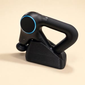 Op de afbeelding: Een zwart draagbaar massageapparaat met een afgeronde kop en een handvat, rustend op een zwarte oplaadbasis. Het apparaat heeft een ronde knop met een blauw licht. Het algehele ontwerp is strak en modern.