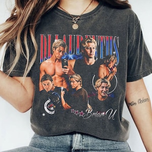 May include: Dark gray t-shirt featuring a collage of images of a man, with the name "Brian U" and the word "Laurentius" in a retro style. The design includes red, blue, and white accents, and a circular emblem.