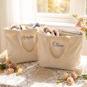 Può includere: Due borse tote in tela beige con manici, una con la scritta "Amelia" e l'altra "Olivia" in corsivo blu. Le borse sono piene di sciarpe e libri. La scena è ambientata su un tavolo con candele e fiori.