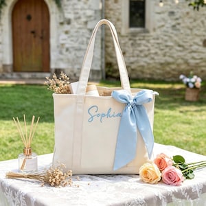Può includere: Borsa tote color crema con il nome "Sophia" ricamato in azzurro. Un nastro azzurro è legato al manico. La borsa è piena di oggetti e si trova su un tavolo con fiori e un diffusore.
