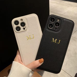 May include: Two smartphone cases with a textured finish, one in cream and one in black. Each case features a triple camera cutout and the gold-colored embossed initials M.J centered on the back. The cases are designed for a modern mobile device.