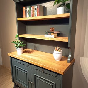 Könnte beinhalten: Ein dunkelgrüner Schrank mit einer Arbeitsplatte aus Naturholz und zwei offenen Regalen. Der Unterbau verfügt über zwei Schranktüren mit Kassettenfronten und silbernen Knöpfen. Die Regale enthalten Bücher, eine kleine Grünpflanze und ein Gefäß.