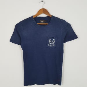 May include: A dark blue short-sleeved t-shirt with a crew neckline and a single chest pocket. The pocket features a white embroidered logo with the text RRL and Double RL surrounded by a laurel wreath design. The shirt is displayed on a wooden hanger.