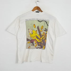 May include: A white short-sleeve t-shirt featuring a graphic print from The Crab with the Golden Claws. The illustration shows a character in a yellow jacket amidst flying gold-colored key-shaped tokens and wooden planks, with the title text above.