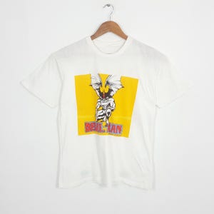 May include: A white short-sleeve t-shirt featuring a central yellow square graphic. The graphic depicts a winged, muscular character in a crouching pose with the red and blue text "DEVILMAN" printed below the figure on the shirt front.