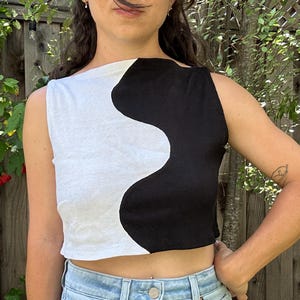 Könnte beinhalten: Ein ärmelloses bauchfreies Oberteil mit einem markanten, vertikalen Yin-Yang-inspirierten Design, bei dem eine Seite in Weiß und die andere in Schwarz gehalten ist. Das Kleidungsstück hat einen geraden Ausschnitt und einen kurzen Saum.
