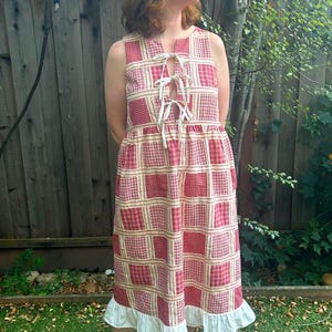 May include: A sleeveless midi dress featuring a red and white checkered grid pattern. The bodice includes a front slit with two white fabric ties for closure. The skirt has a gathered waist and a solid white ruffled hem at the bottom.