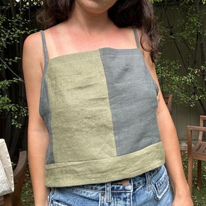 May include: A sleeveless square-neck top featuring a color-blocked design with a vertical split of olive green and charcoal gray fabric. The garment has thin shoulder straps and a horizontal band detail at the bottom hem, constructed from a textured material.