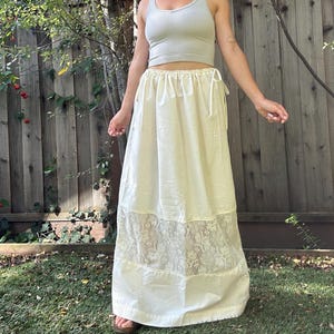 May include: A light gray ribbed tank top and a long cream-colored skirt. The skirt features an elasticized drawstring waistband and a wide horizontal band of intricate floral lace detailing positioned near the bottom hem of the garment.