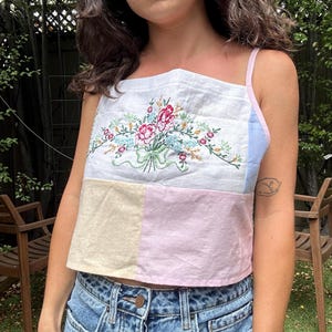 May include: A sleeveless crop top featuring a white upper panel with a colorful floral embroidery design, a light blue side panel, and a lower section color-blocked with beige and light pink fabric panels, finished with thin pink shoulder straps.