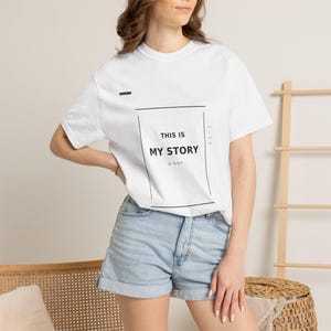 May include: White t-shirt with the text "THIS IS MY STORY" in a black rectangular frame. The shirt also has additional text in a smaller font. The model is wearing denim shorts.