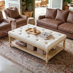 May include: A rectangular tufted ottoman with a cream-colored fabric top and a light wood frame. The piece features a lower shelf holding two woven baskets and books. A wooden tray with a candle and books sits on the tufted surface.