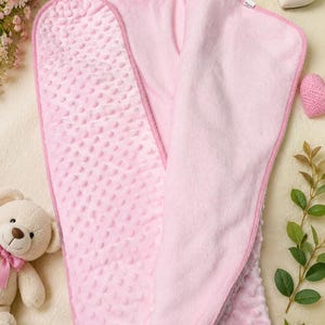 May include: A soft, light pink baby blanket featuring a textured dimpled minky fabric on one side and a smooth, solid pink fabric on the reverse. The blanket is rectangular with rounded corners and finished with a matching pink fabric trim edge.
