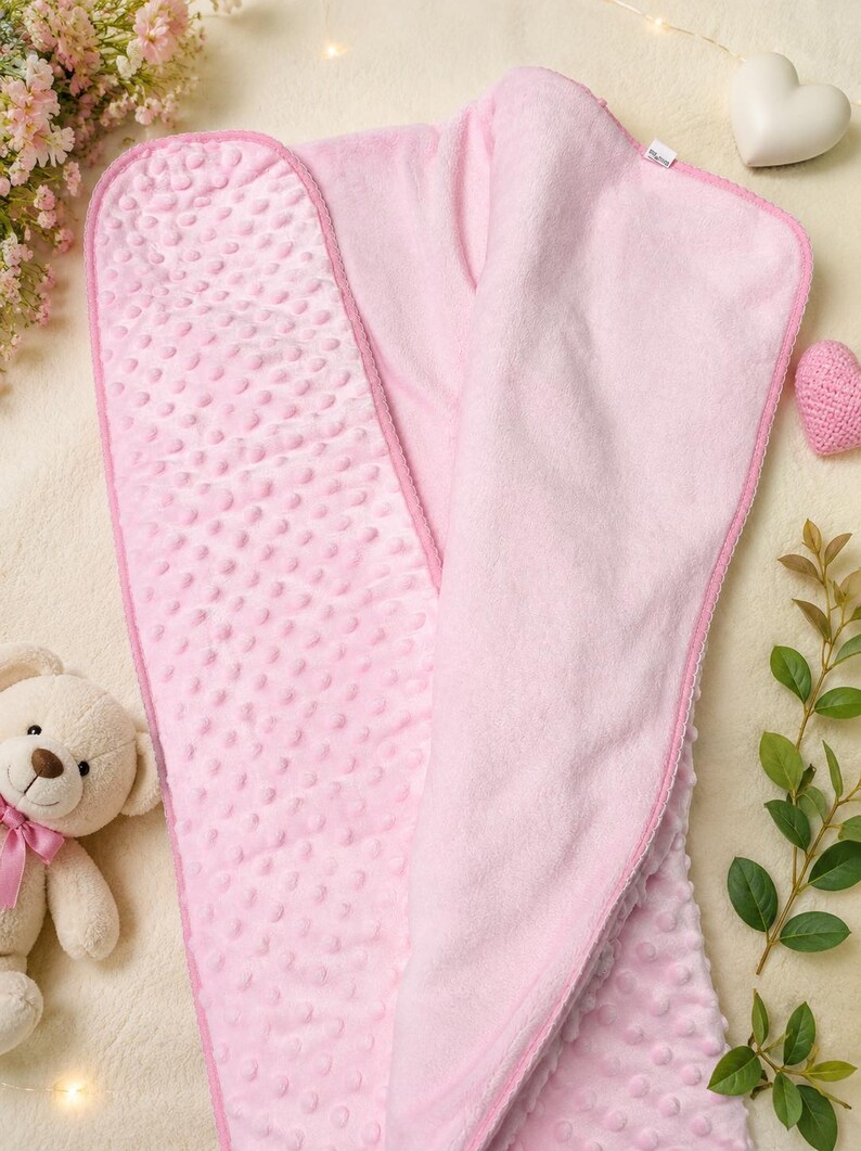 May include: A soft, light pink baby blanket featuring a textured dimpled minky fabric on one side and a smooth, solid pink fabric on the reverse. The blanket is rectangular with rounded corners and finished with a matching pink fabric trim edge.