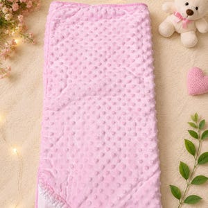 May include: A rectangular baby blanket featuring a soft light pink minky fabric with a raised dimple texture. The blanket has a folded corner revealing a white mesh lining and a decorative white lace trim along the edge of the fabric fold.