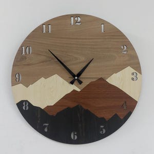 May include: A round wooden wall clock featuring a layered mountain landscape design in shades of light beige, medium brown, and dark charcoal. The clock face includes cut-out numerals from 1 to 12 and two black hands at the center.