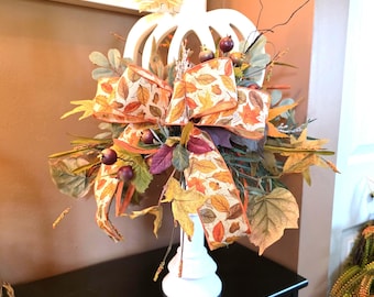 May include: A white wooden pumpkin cutout centerpiece mounted on a pedestal base, adorned with a large fabric bow featuring a colorful autumn leaf pattern, surrounded by artificial fall foliage, dark berries, and thin twigs in shades of orange and brown.