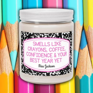 Pode incluir: Uma vela de pote branca com tampa branca e um rótulo que diz "Smells Like Crayons, Coffee, Confidence & Your Best Year Yet, Miss Jackson." O fundo apresenta lápis coloridos.