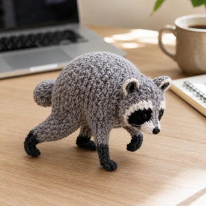 May include: A crocheted raccoon figurine crafted from grey yarn with a white muzzle, black eye patches, and dark grey paws. The small stuffed animal is depicted in a walking pose with a rounded back, featuring a black nose and small ears.