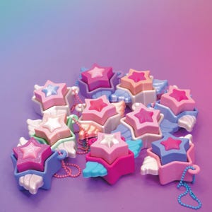 May include: A collection of star-shaped pendants in various shades of pink, blue, and white. Each pendant features a smaller star on top and decorative wings. Some have beaded chains attached.