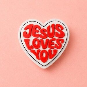 May include: A heart-shaped charm featuring a white base with a black outline and the raised red text "JESUS LOVES YOU" in a bold, rounded, bubble-style font. The design is centered on the heart, with the words stacked in three distinct lines.