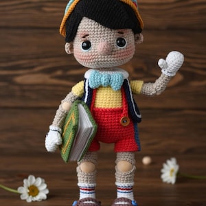 May include: A handmade crochet doll of Pinocchio, wearing a yellow shirt, red shorts, and a blue vest. The doll holds a green book and has a blue bow tie. The doll is wearing a hat and white gloves.